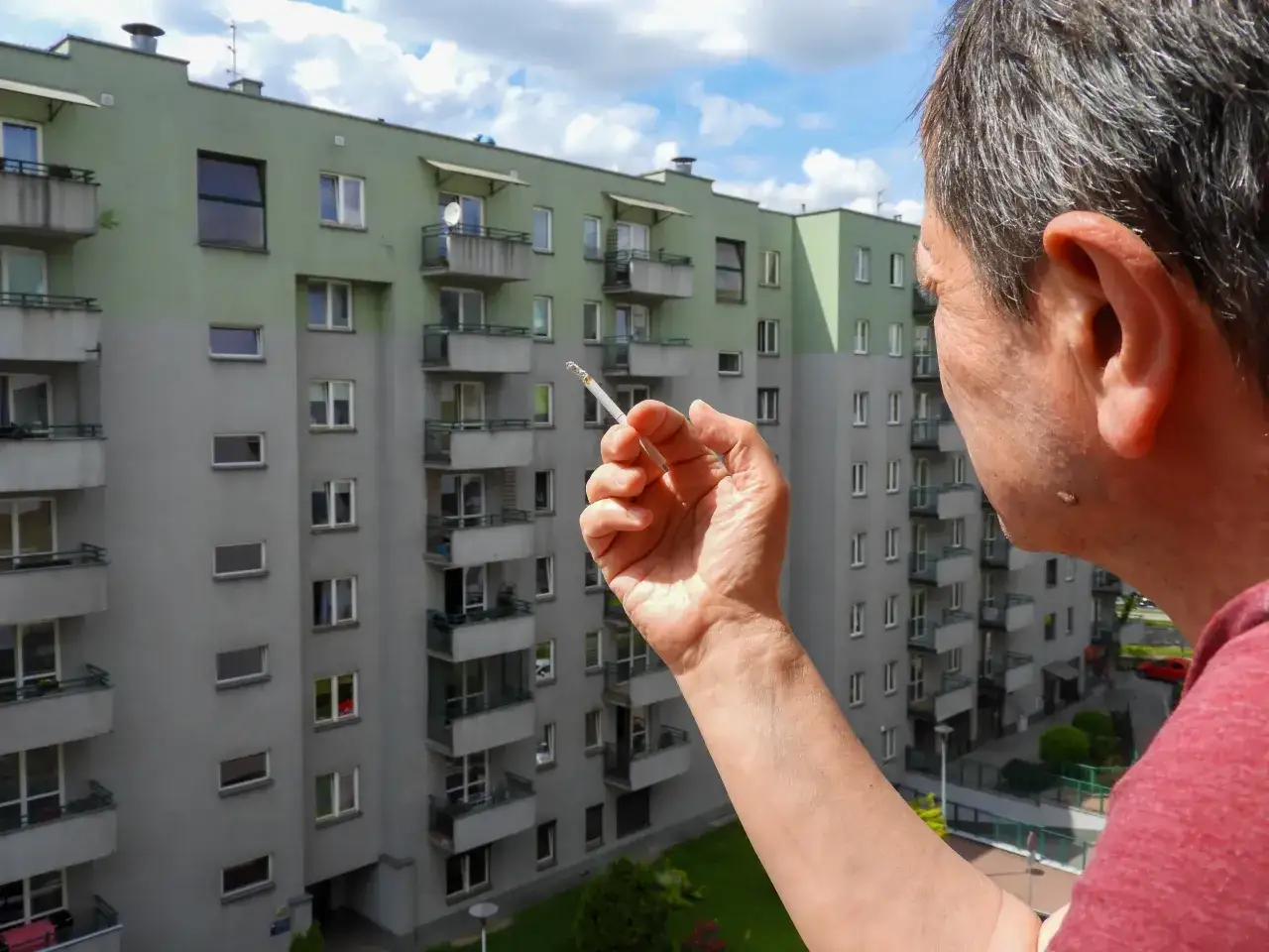 Mężczyzna trzyma papierosa, zastanawiając się, czy można palić na balkonie. W tle blok mieszkalny.