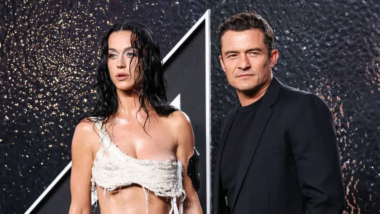 Katy Perry i Orlando Bloom na tle błyszczącej ściany. Katy w odważnej, postrzępionej kreacji, Orlando w eleganckim garniturze.