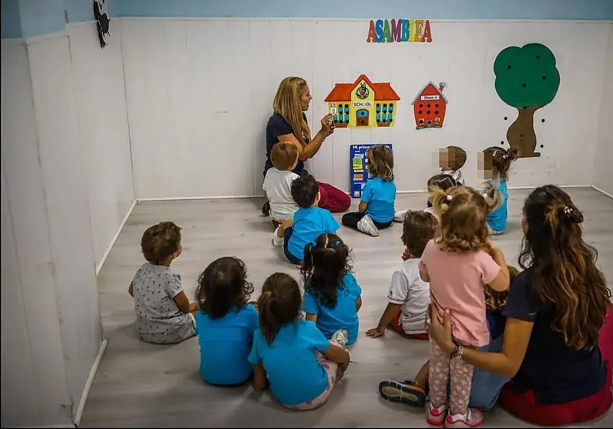 Maestra ense&ntilde;a a ni&ntilde;os de guarder&iacute;a de 2 a&ntilde;os sobre la escuela. Decoraci&oacute;n de pared con casas y &aacute;rbol.