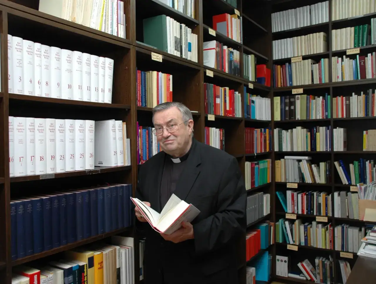 Kardinal Lehmann in seiner Bibliothek, umgeben von B&uuml;chern. Er h&auml;lt ein Buch in der Hand.