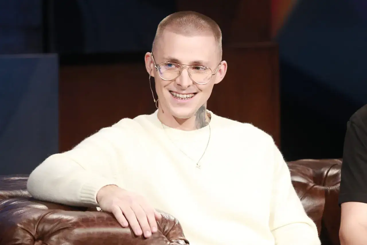 Młody mężczyzna z tatuażami na szyi, w okularach i swetrze, uśmiecha się. Mateusz Trąbka wiek nie jest widoczny, ale wygląda na około 20 lat.