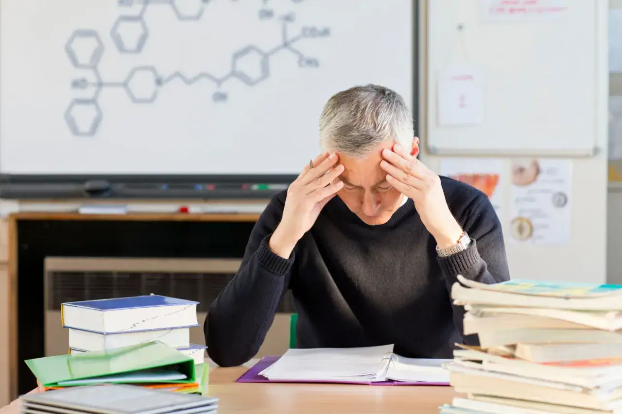 Lehrer mit H&auml;nden an den Schl&auml;fen, &uuml;berfordert von der Arbeit. Die chemische Formel an der Tafel deutet auf seine Belastung hin, ein Zeichen von Lehrer Depression.