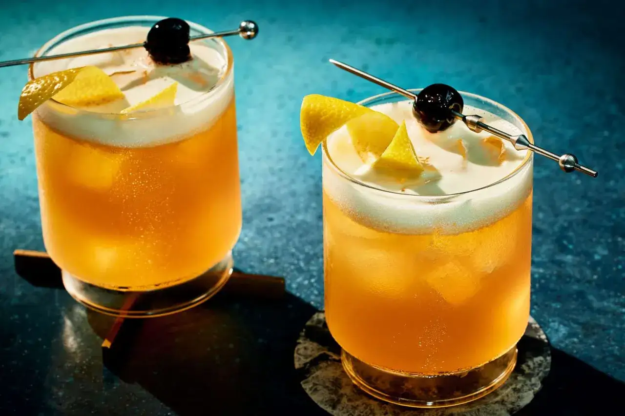 Zwei erfrischende Amaretto Sours mit Schaumkrone, Kirsche und Zitronenzeste auf Eis.