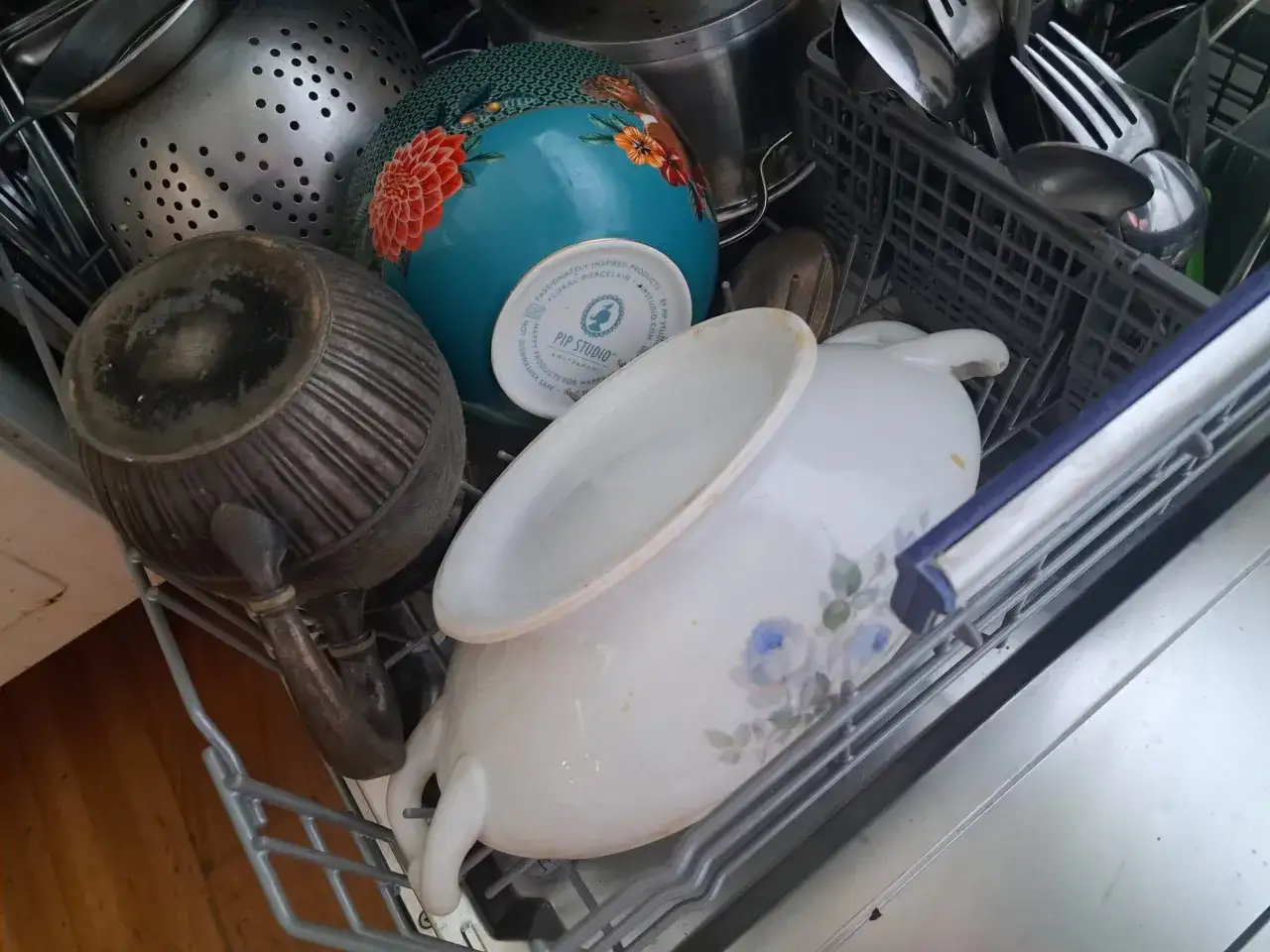 Stare naczynia, w tym metalowy dzbanek i porcelanowa miska z kwiatami, w zmywarce. Na spodzie miski napis 