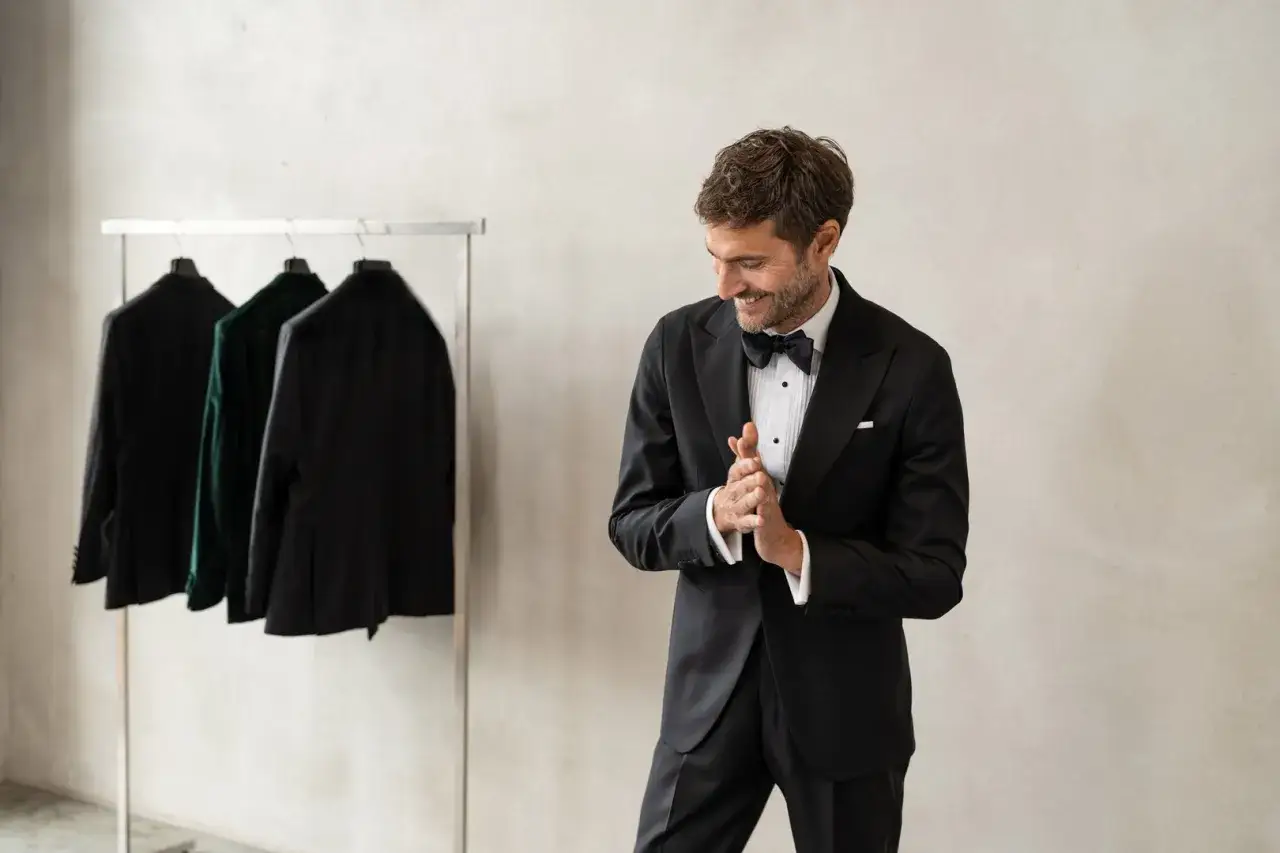 Man i smoking redo för black tie dresscode. Tre kavajer hänger på en klädställning i bakgrunden.