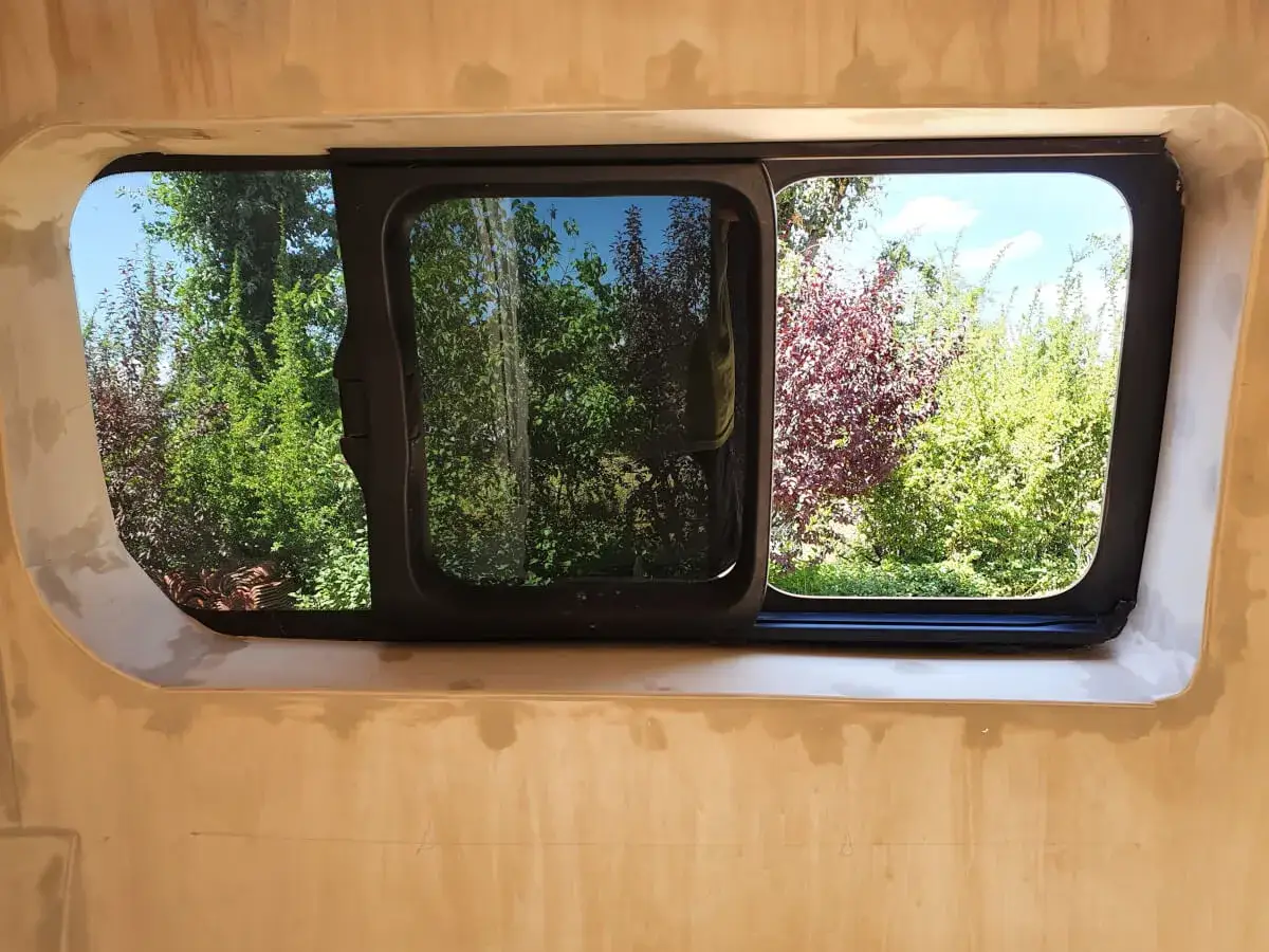 Fenêtre de fourgon aménagé, prête pour le contrôle technique. Vue sur un jardin verdoyant.