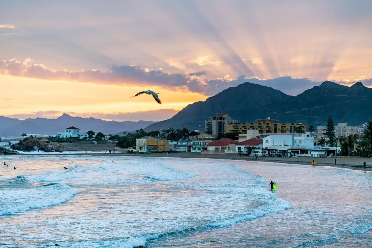 Atardecer en las playas de Mazarrón. Un ave surca el cielo mientras las olas acarician la costa y los surfistas disfrutan del mar.
