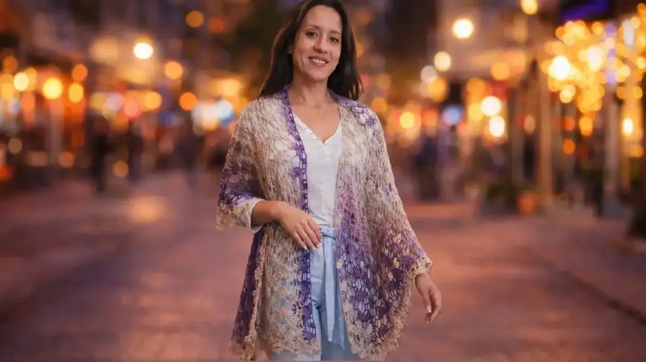 Mujer sonriente luce un chal rectangular tejido a mano, con degradado de morado a beige, sobre un fondo de luces urbanas nocturnas.
