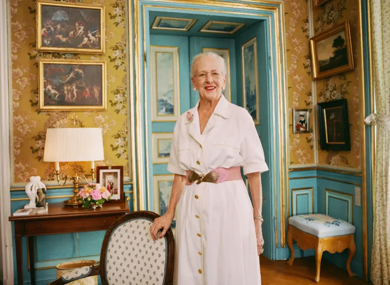 Äldre dam i vit klänning med rosa bälte, står i ett rum med gulblommig tapet och turkosa detaljer. Hon ser ut att må bra, trots att man kan tro att drottning Margrethe är sjuk.