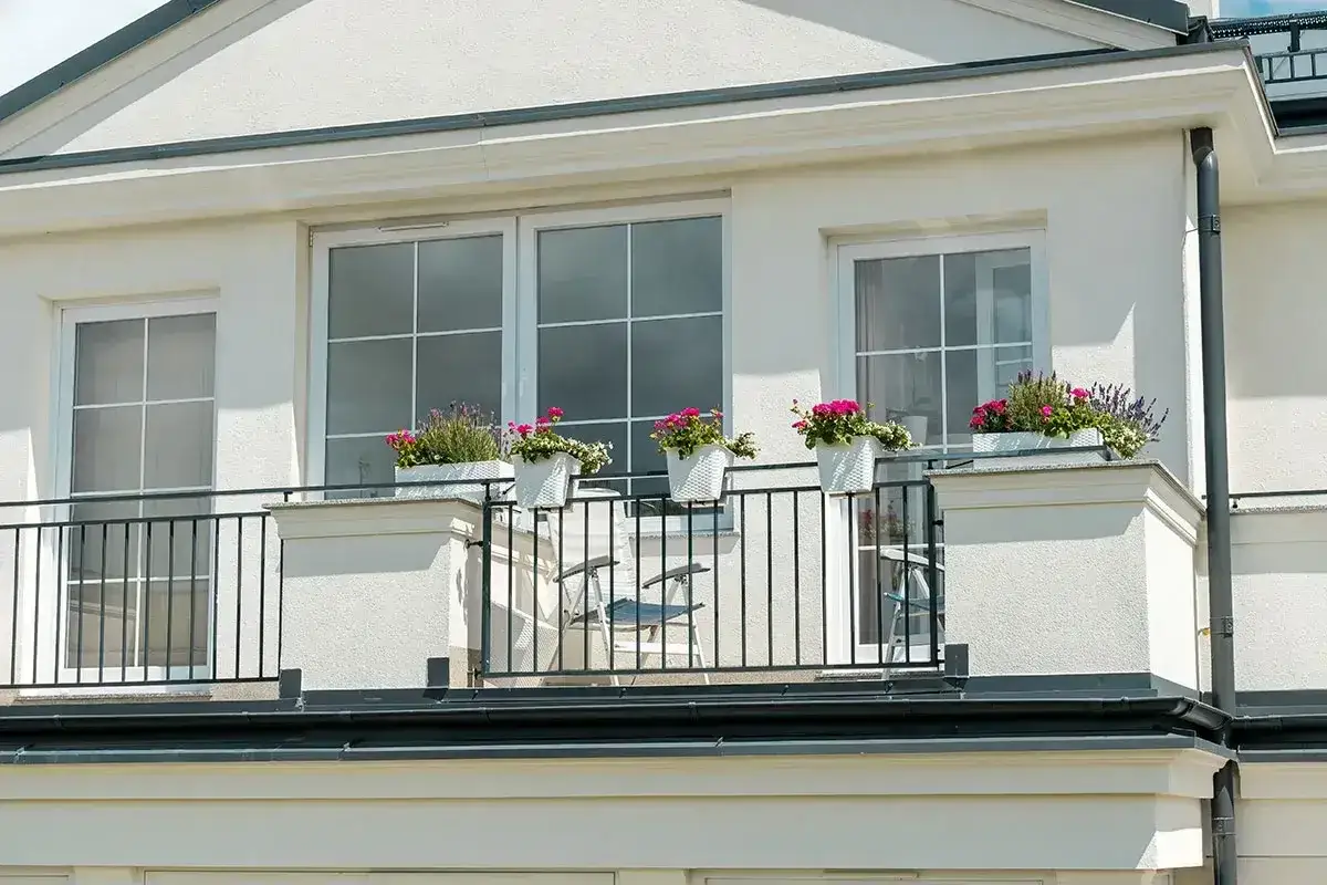 Białe okna pięknie komponują się z jasną, tynkowaną elewacją, tworząc elegancki wygląd. Balkon ozdobiony kwiatami dodaje uroku.