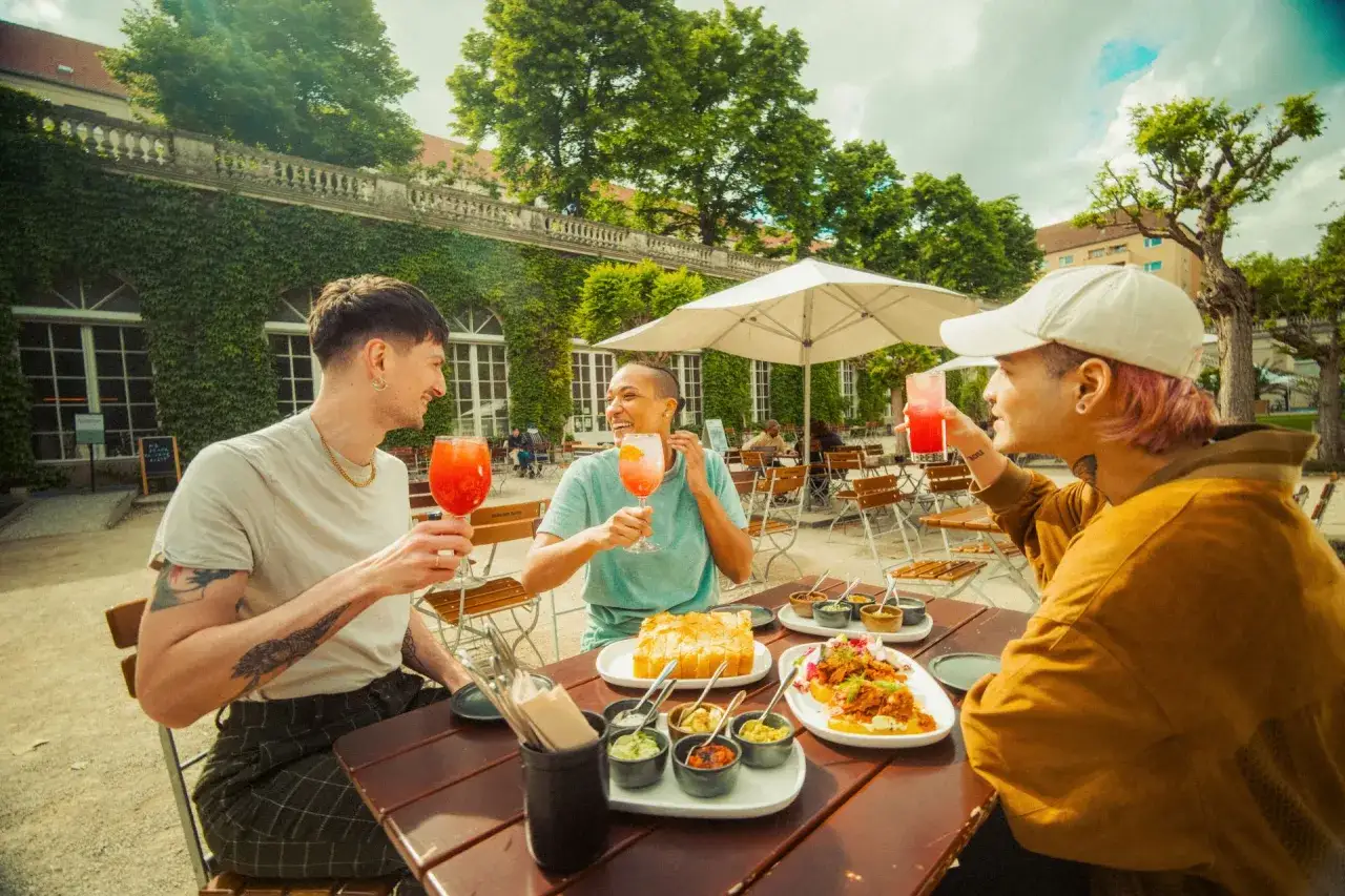 Drei Freunde genie&szlig;en Aperol Spritz und leckeres Essen in einem charmanten Biergarten, ein perfekter Ort f&uuml;r Berlin Foodspots.