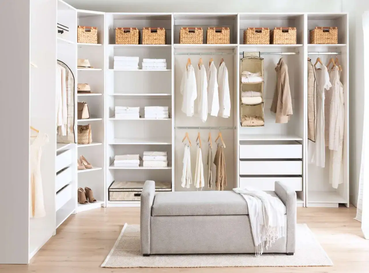 Armario blanco con cestas de mimbre, ropa colgada y doblada, y un banco. Ideas para organizar un armario sin cajones.