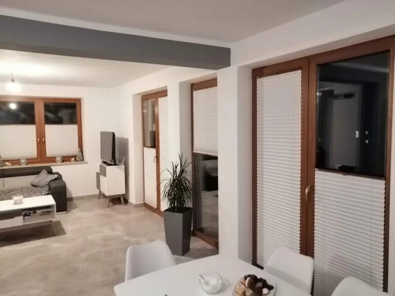 Nowoczesny salon z białymi plisy na oknach i drzwiach balkonowych. Eleganckie rozwiązanie, które zastępuje tradycyjne rolety.