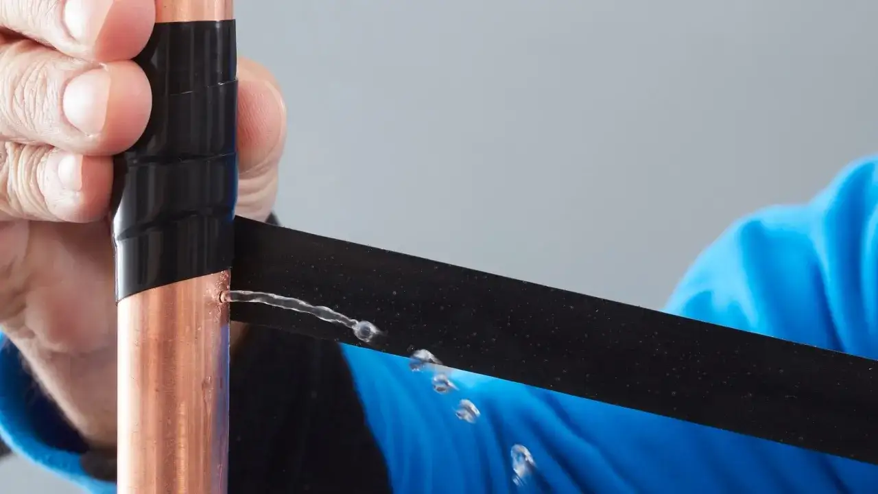 Hand dichtet Kupferrohr mit schwarzem Klebeband ab, um einen Wasseranschluss abzudichten. Wasser spritzt aus einem Loch.