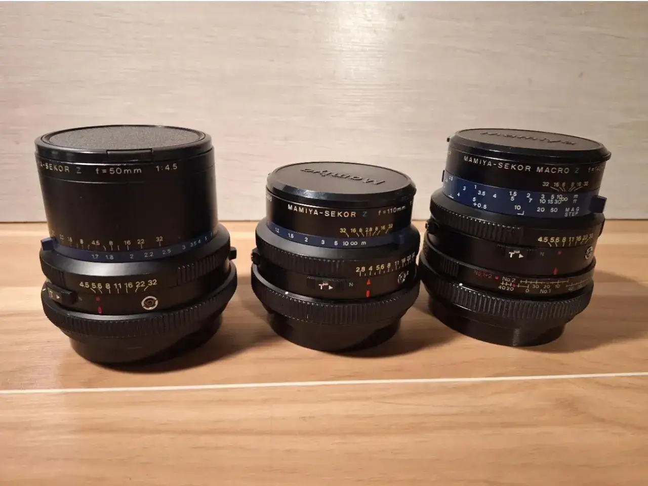 Three Mamiya-Sekor Z lenses, including a 50mm f/4.5, 110mm f/2.8, and a macro lens, sit on a wooden surface.