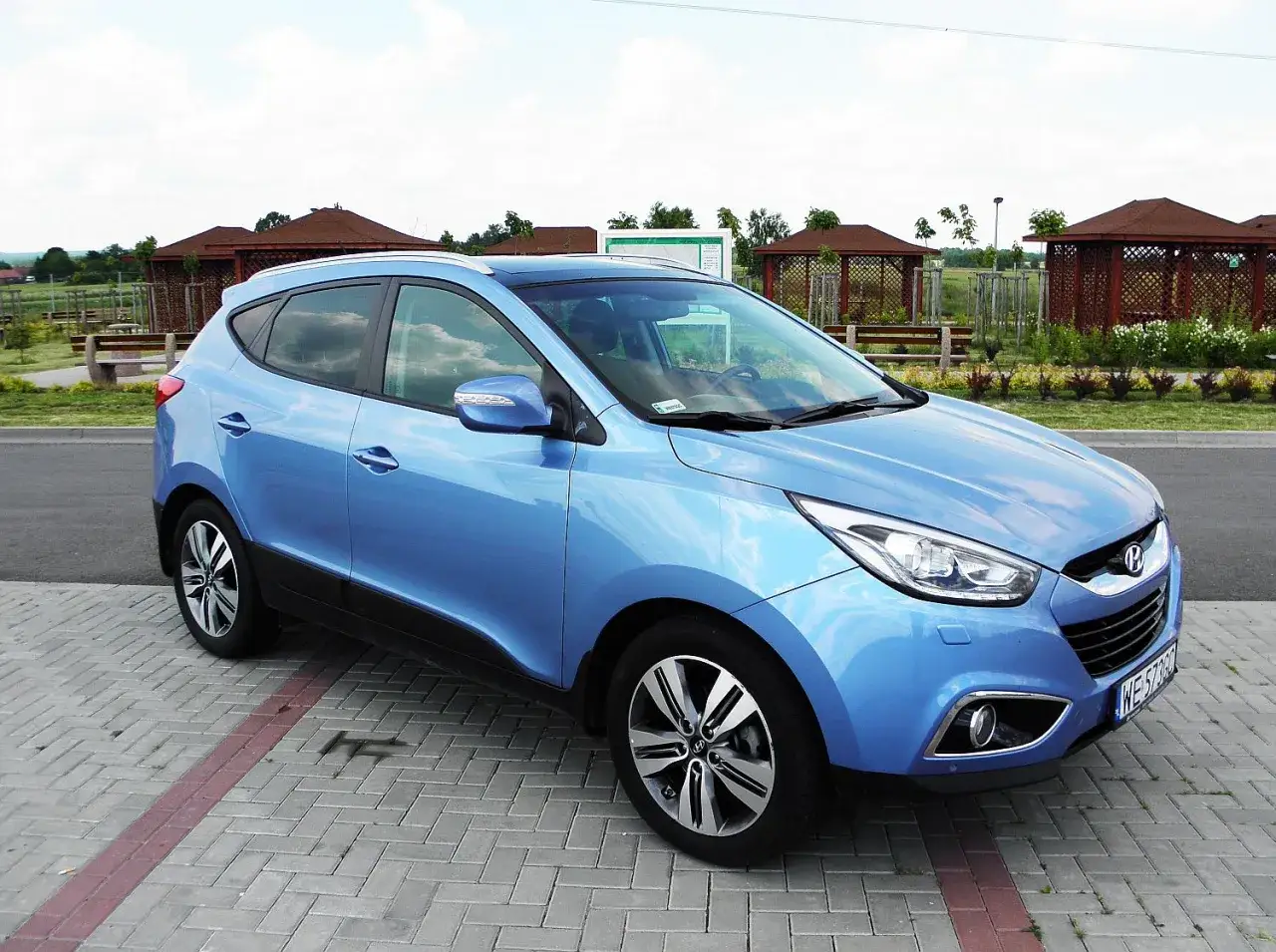 Błękitny Hyundai ix35 2.0 benzyna zaparkowany na wybrukowanym placu, w tle altanki i zieleń.