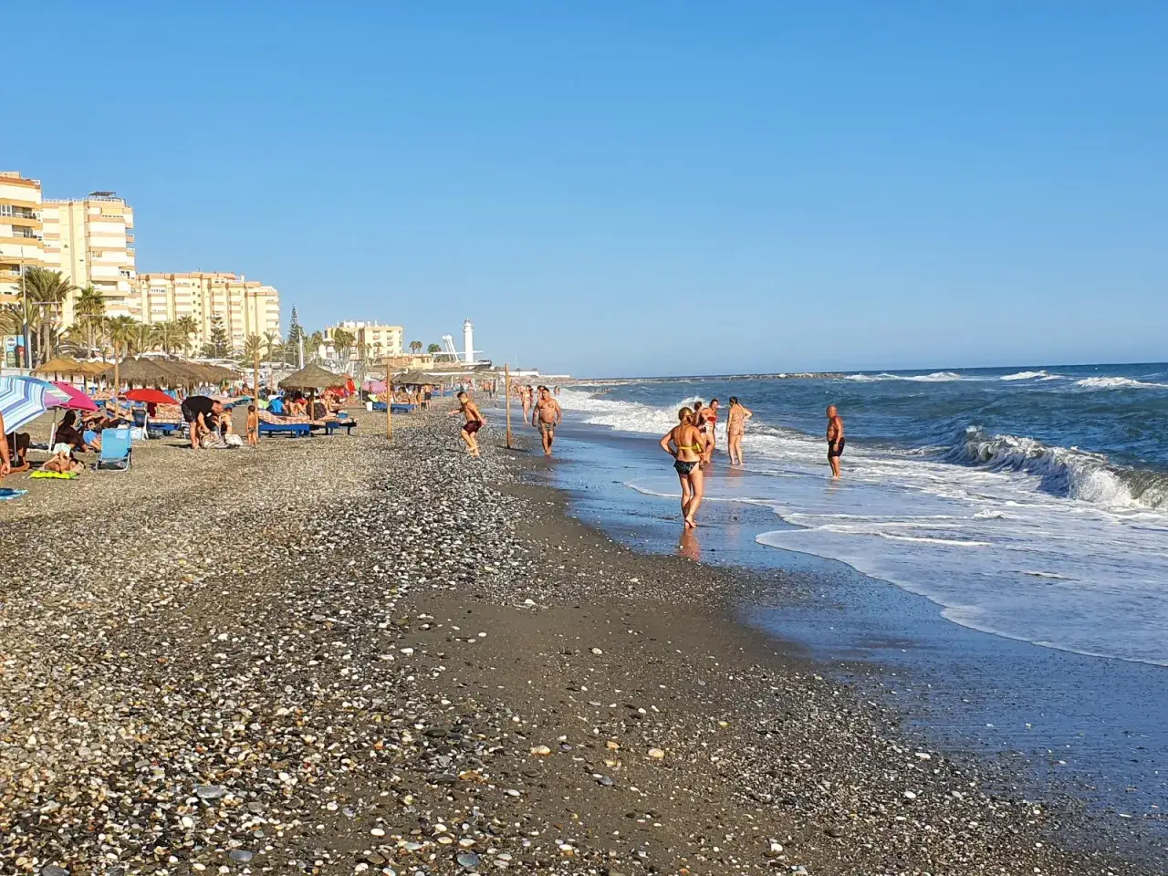 Menschen genie&szlig;en einen sonnigen Tag am torrox strand. Wellen brechen am Kiesstrand, w&auml;hrend im Hintergrund Geb&auml;ude und ein Leuchtturm zu sehen sind.