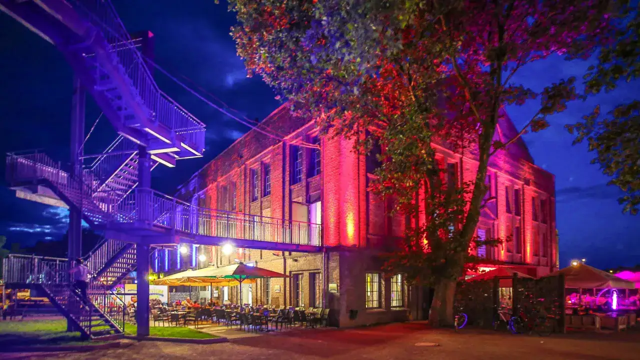 Abendstimmung im **Extraschicht Dorsten**: Eine beleuchtete Treppe führt zu einem Gebäude, das in rotes Licht getaucht ist. Draußen laden Tische und Stühle zum Verweilen ein.