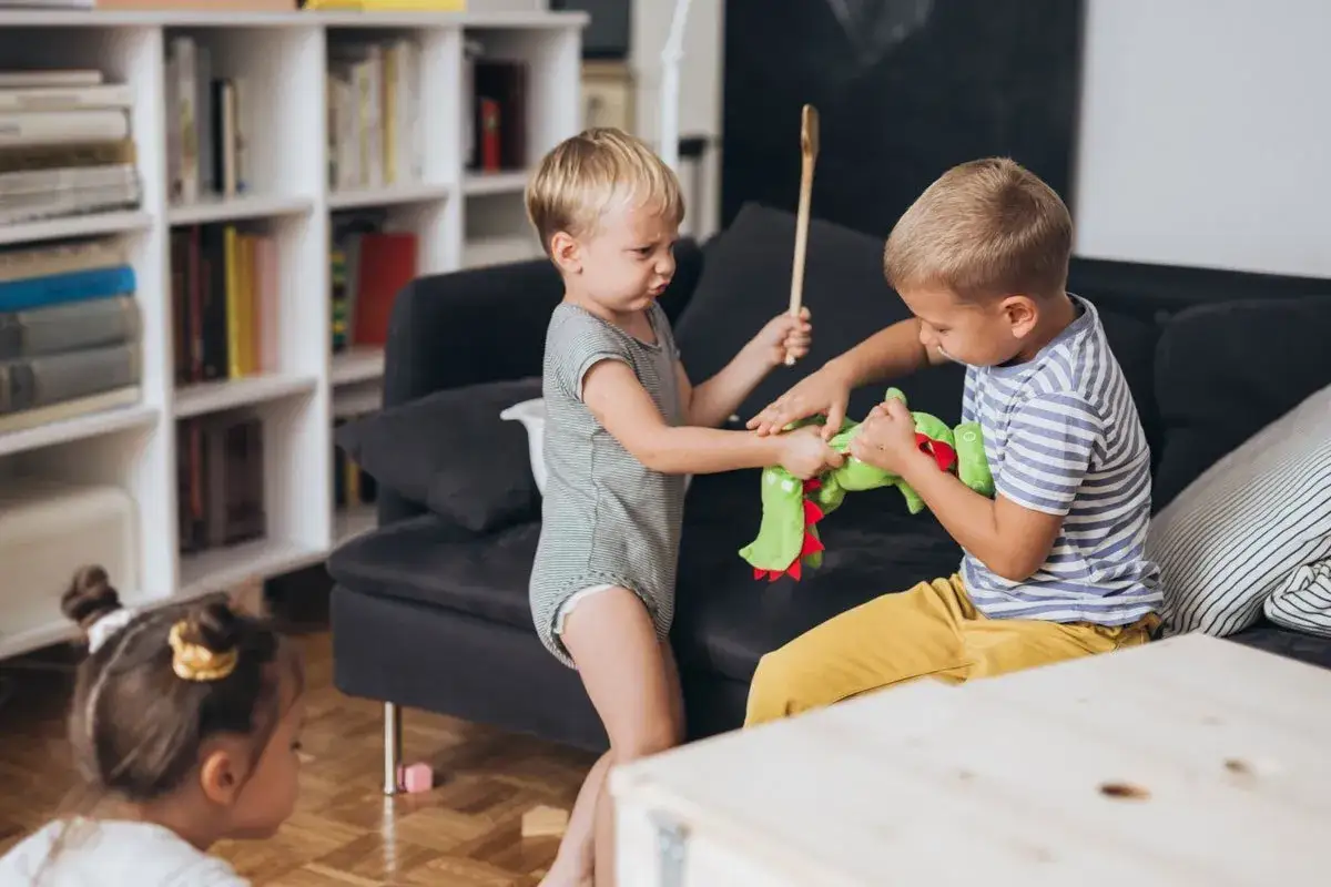 Zwei kleine Jungen streiten sich um ein grünes Stofftier. Einer hält einen Kochlöffel, der andere zieht am Spielzeug. Ein kleines Kind schaut zu.