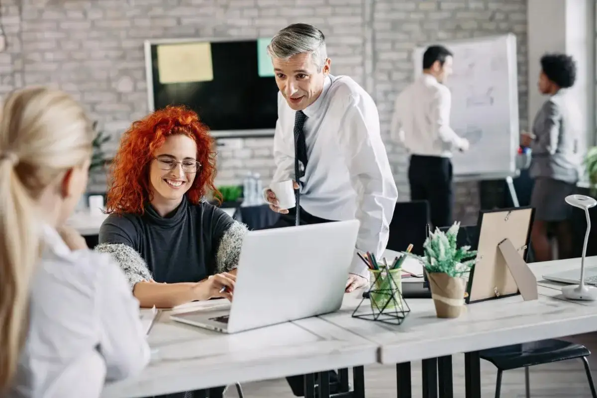 Pracownicy w biurze dyskutują przy laptopie. W tle mężczyzna pisze na tablicy. Czy pracodawca może nagrywać pracowników?