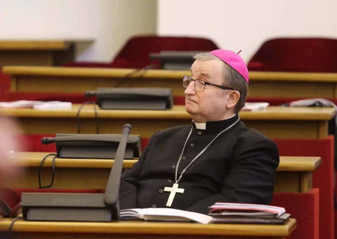 Biskup w stroju duchownym, z różowym piuską i krzyżem na łańcuchu, siedzi w sali konferencyjnej. Czy biskup to zawód? Tak, to powołanie i służba.