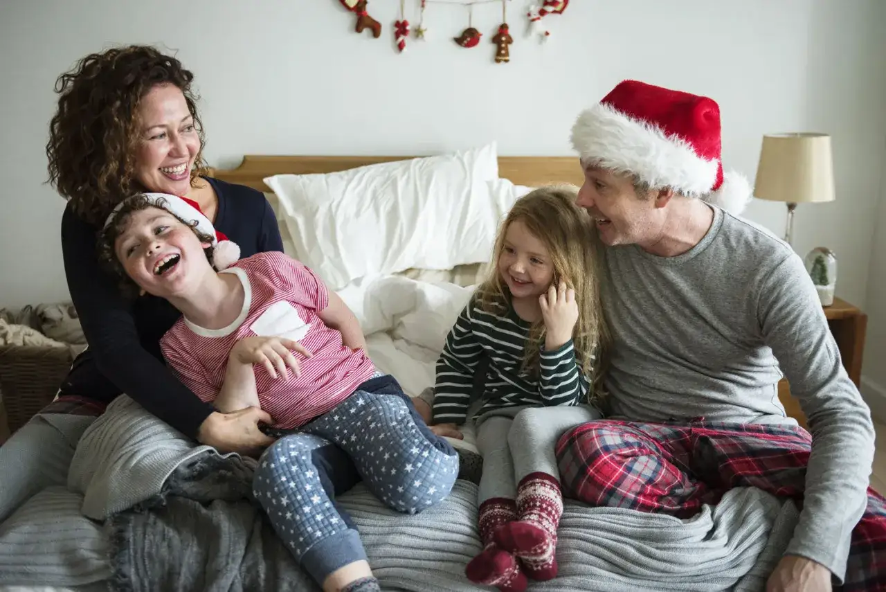 Rodzina w świątecznych piżamach śmieje się na łóżku. To kwintesencja parenting co to – wspólne, radosne chwile.