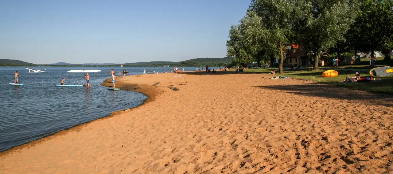 Sonniger Strand am Niedernberger See mit Menschen beim Stand-Up-Paddling und Entspannen.