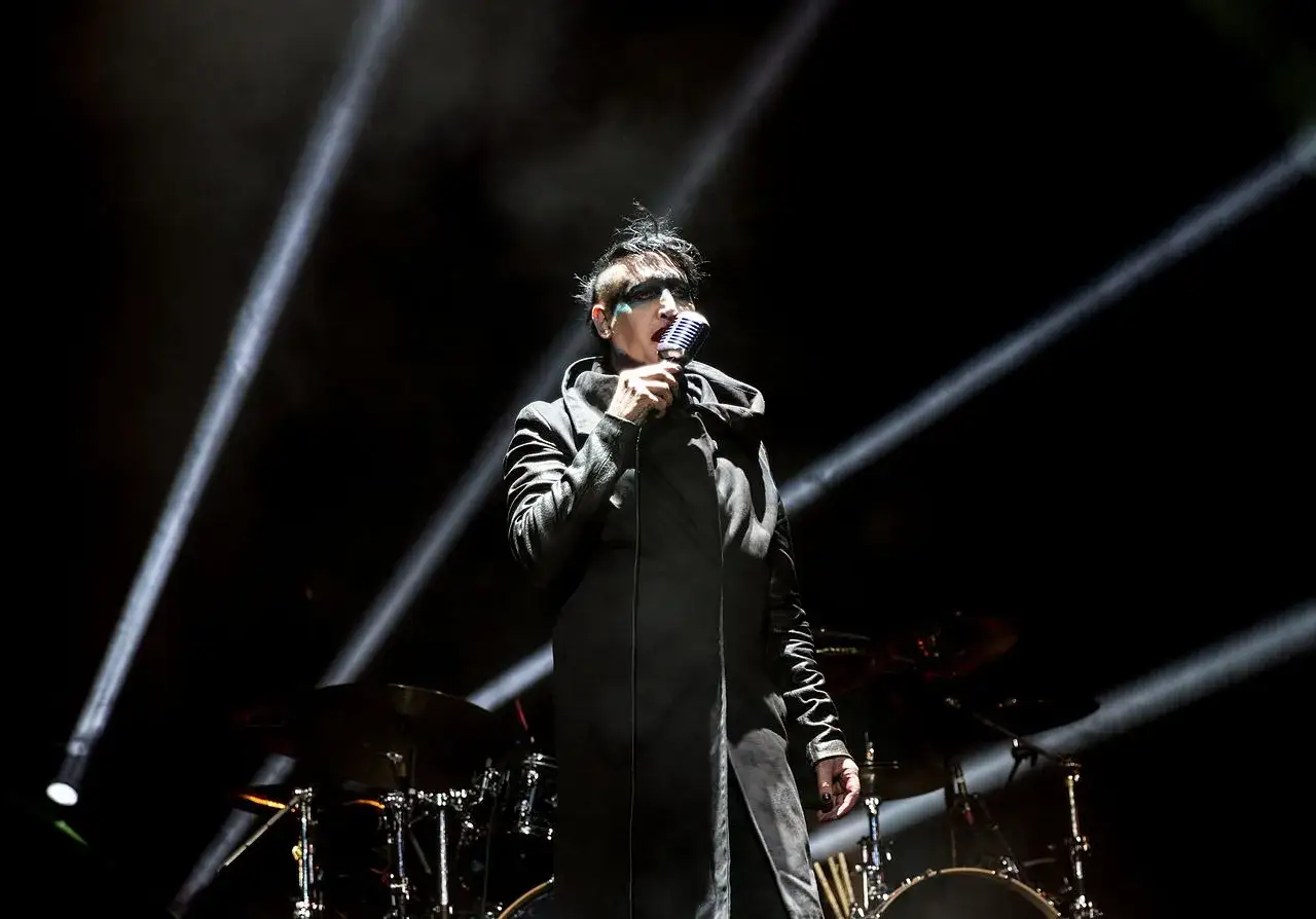 Marilyn Manson na koncercie w Warszawie, w czarnym płaszczu i z charakterystycznym makijażem, śpiewa do mikrofonu.