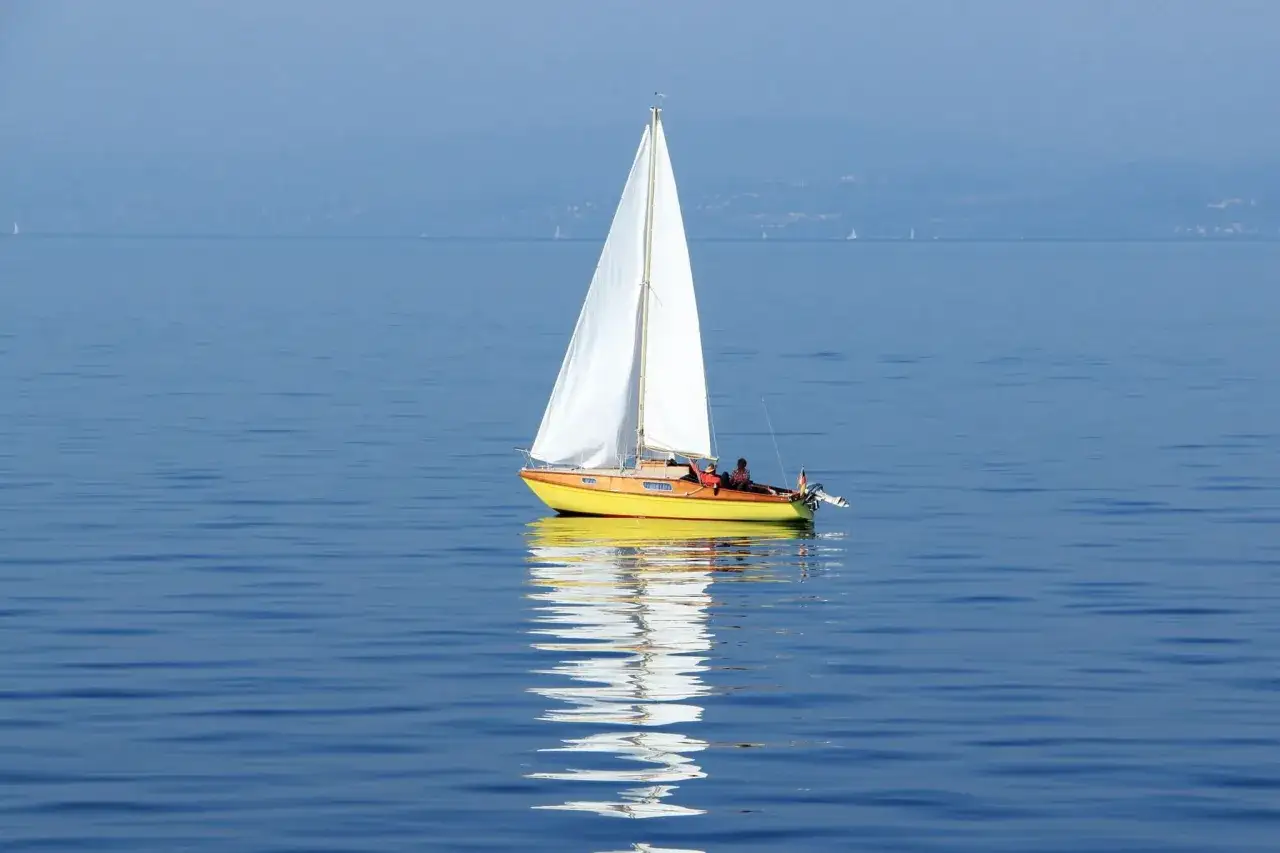 Ein gelbes Segelboot mit weißem Segel gleitet auf ruhigem Wasser. Im Hintergrund sind weitere kleine Segelboote und eine ferne Küstenlinie zu sehen, die verschiedene kleine Segelboote Typen andeuten.