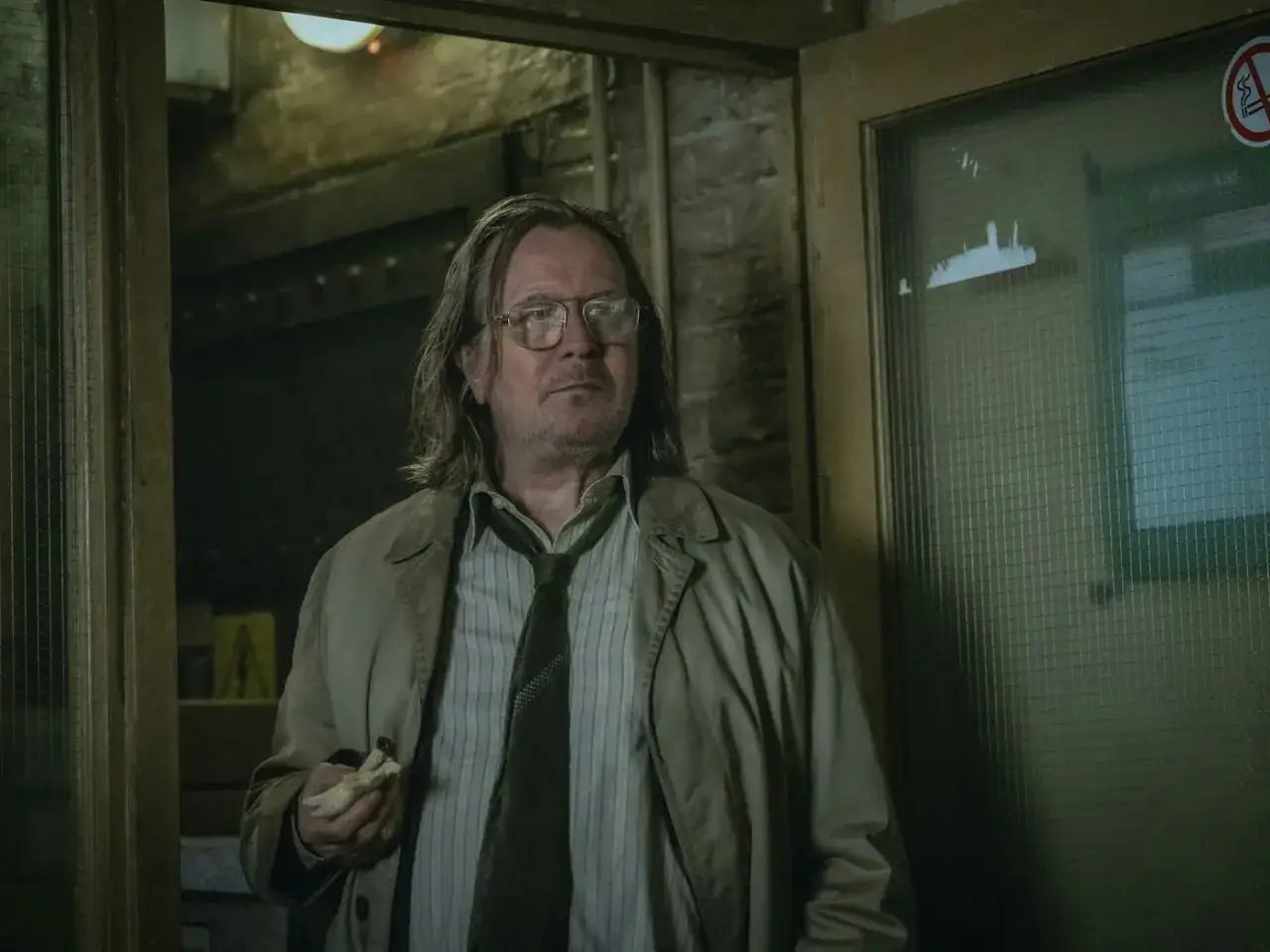 Gary Oldman als Jackson Lamb in slow horses staffel 3, mit zerzaustem Haar und einem Snack in der Hand, blickt nachdenklich in die Ferne.