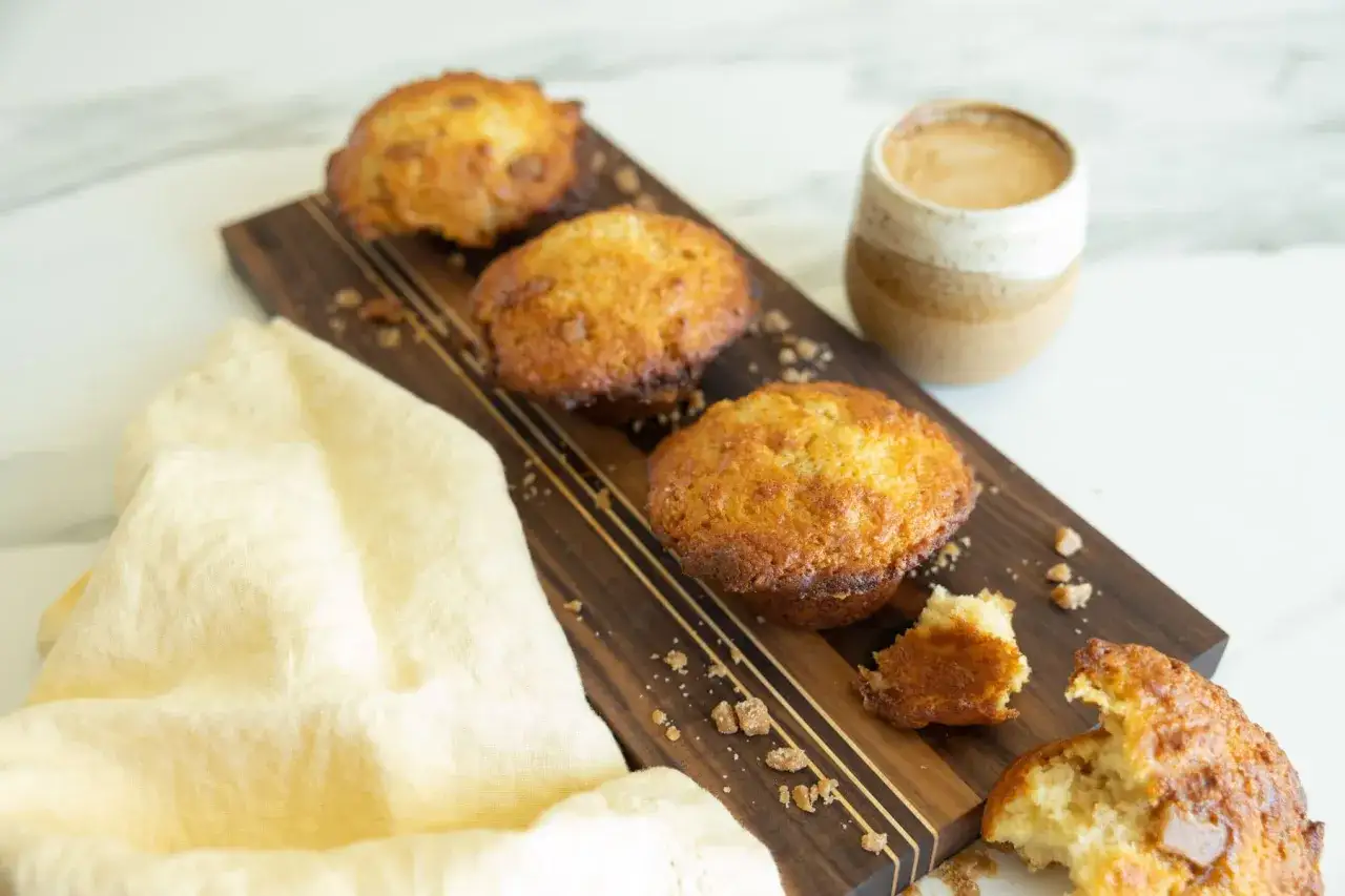 Trois muffins banane caramel dorés sur une planche en bois, avec une tasse de café et un tissu jaune.