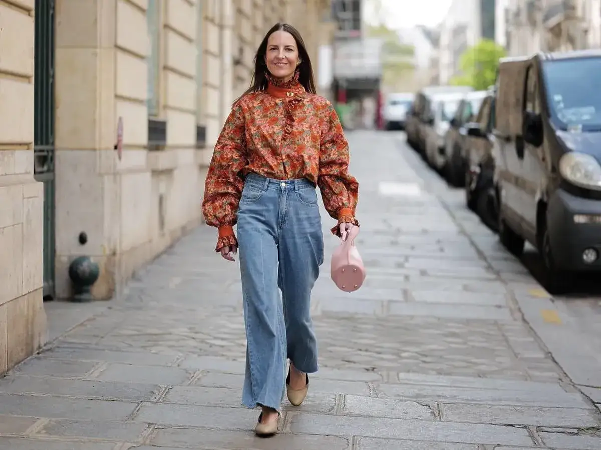 Elegancka figura kolumna w kwiecistej bluzce i jeansach spaceruje po paryskiej ulicy, niosąc różową torebkę.