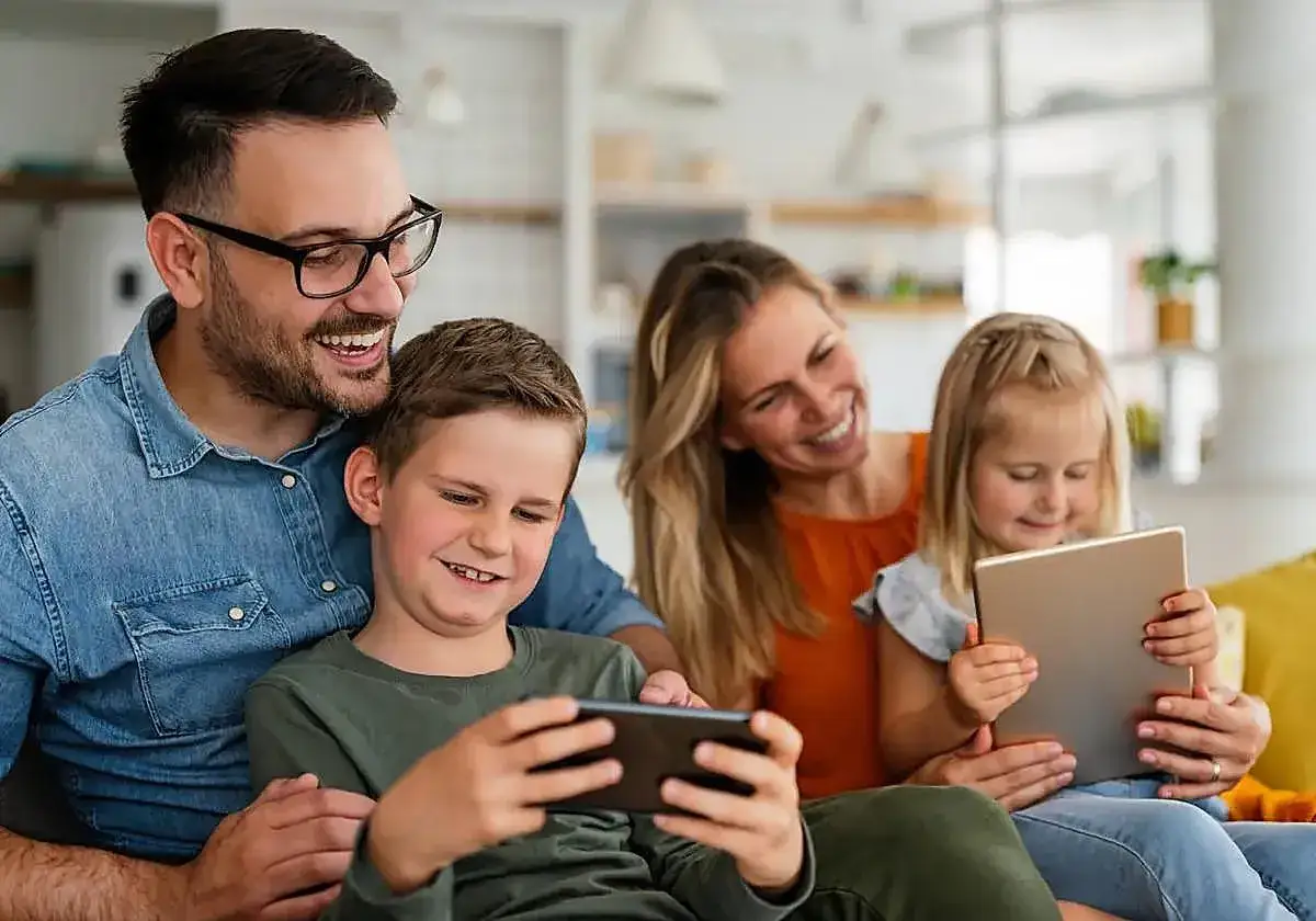 Familia sonriente compartiendo momentos frente a pantallas. Los ni&ntilde;os disfrutan de sus dispositivos, mientras los padres observan con alegr&iacute;a.