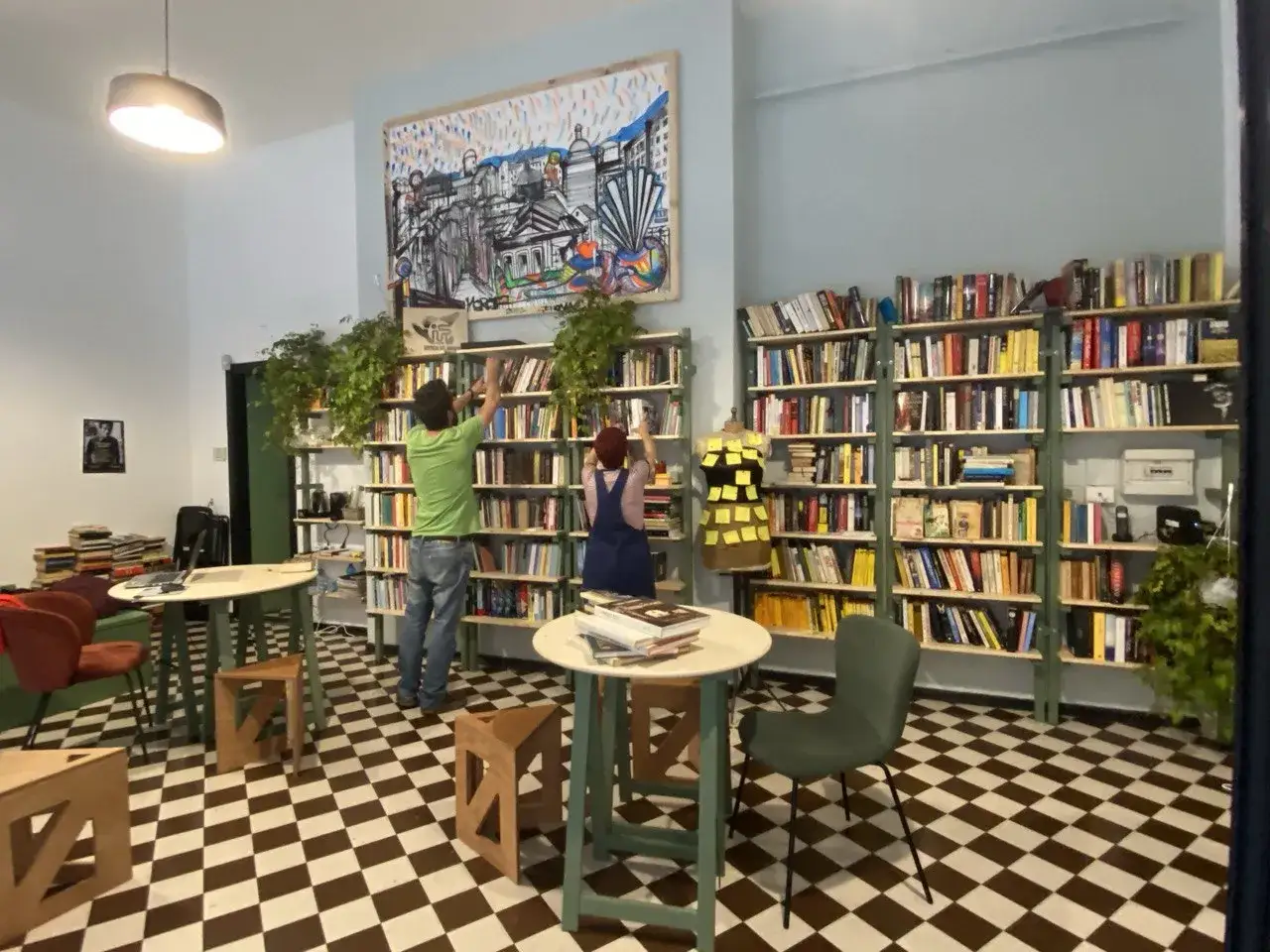 Una **little free library** con scaffali pieni di libri, due persone che sistemano volumi e un'opera d'arte colorata alle pareti.