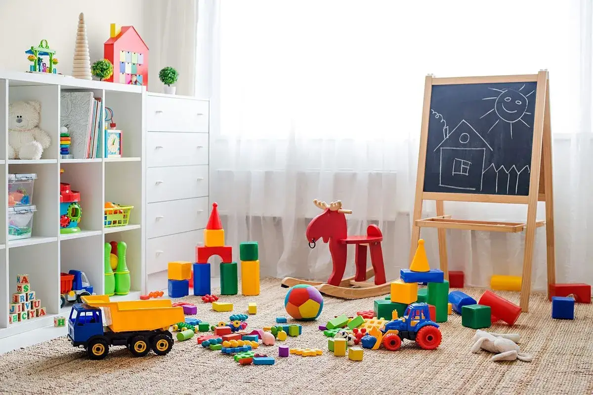 Kolorowy pok&oacute;j zabaw dla dzieci w domu. Na dywanie rozrzucone klocki, ciężar&oacute;wka, traktorek, konik na biegunach i tablica z rysunkiem domu.