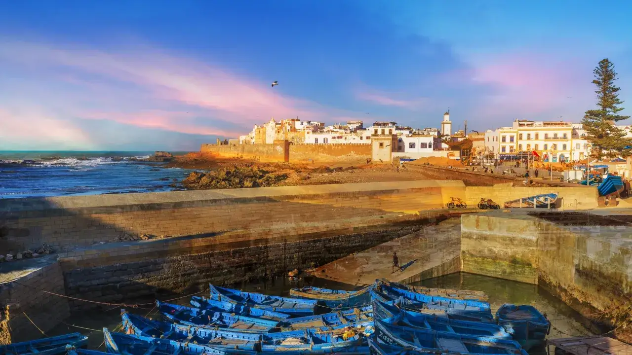 Atardecer en Essaouira: un puerto con barcas azules, murallas doradas y casas blancas bajo un cielo rosado. ¡Essaouira que ver!