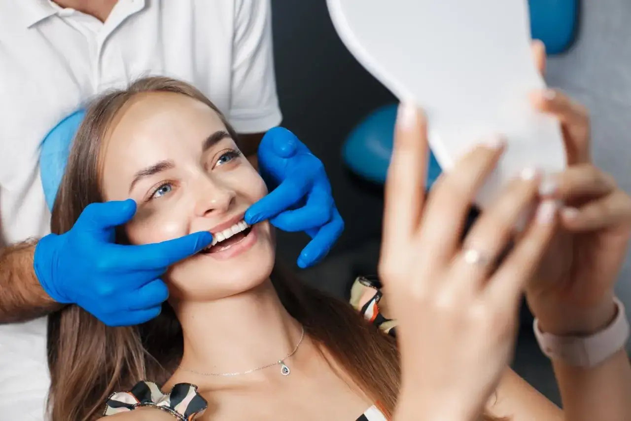 Uśmiechnięta kobieta z niebieskimi rękawiczkami dentysty, która pokazuje jej zęby w lustrze. Proces wybielania zębów.