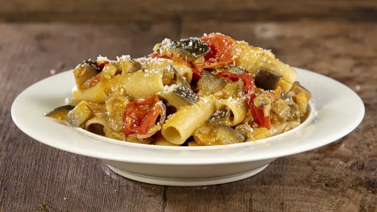 Piatto di pasta con melanzane e pomodorini, condita con formaggio grattugiato, su un tavolo di legno rustico.