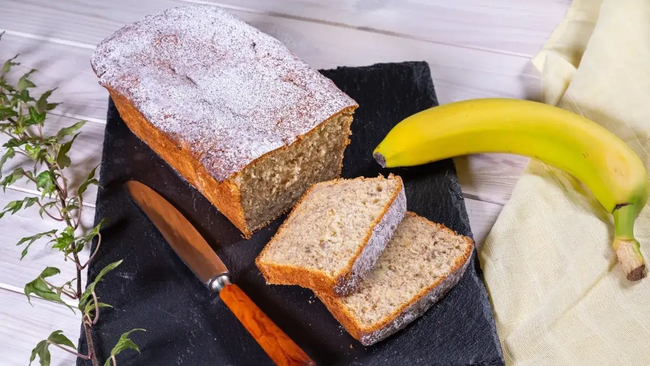 Ein saftiger Bananenkuchen, bestäubt mit Puderzucker, liegt auf einem schwarzen Stein. Daneben ein Messer und eine Banane – perfekt für ein schnelles Bananenkuchen Rezept.