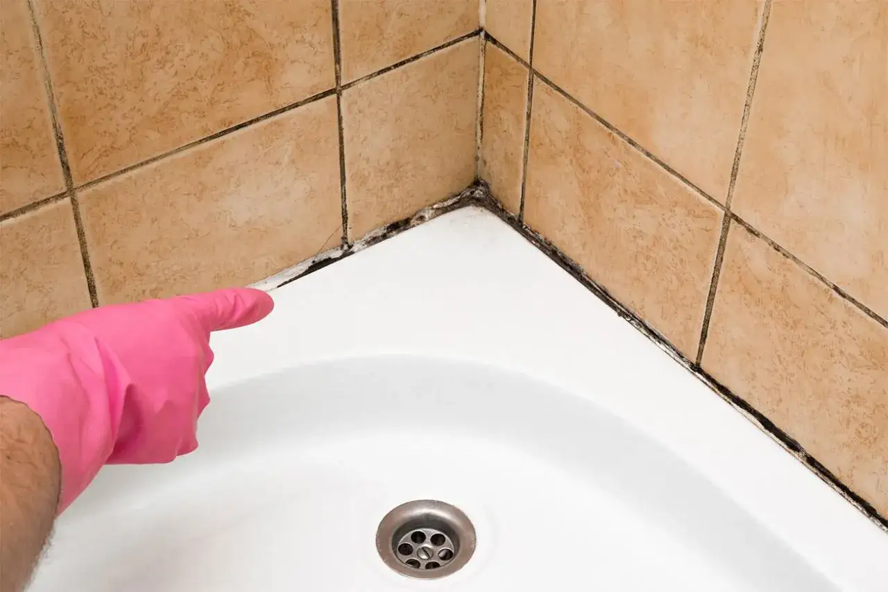 Mano con guanto rosa indica la **muffa in bagno** che si è formata nell'angolo tra le piastrelle e la vasca.