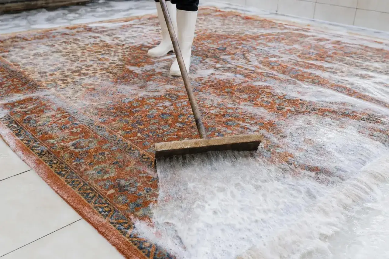 Person mit Gummistiefeln schrubbt mit einem Besen einen orientalischen Teppich. Das Natron hilft beim Teppich reinigen.
