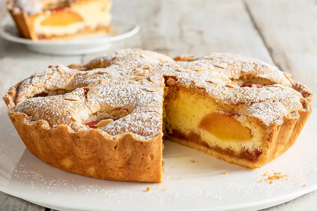 Torta con pesche sciroppate, soffice e dorata, cosparsa di zucchero a velo e mandorle. Una fetta mostra il ripieno succoso.