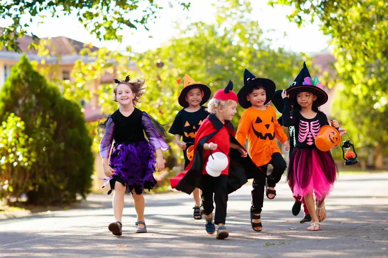 Dzieci w strojach na Halloween, jak zrobić przebranie na Halloween? Wesoła gromadka w dyniowych i szkieletowych kostiumach biegnie po chodniku.