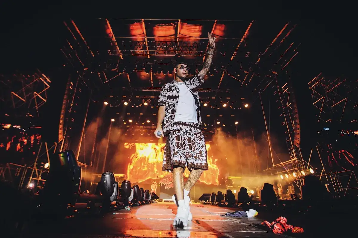 Artista sul palco con un outfit coordinato, luci intense e fumo. Un momento che evoca le canzoni trap pi&ugrave; famose.