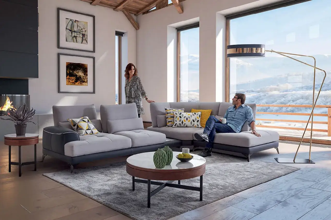 Un couple profite d'un salon moderne avec un canap&eacute; r&eacute;versible, symbole de flexibilit&eacute; et de confort, face &agrave; une vue panoramique sur la montagne.