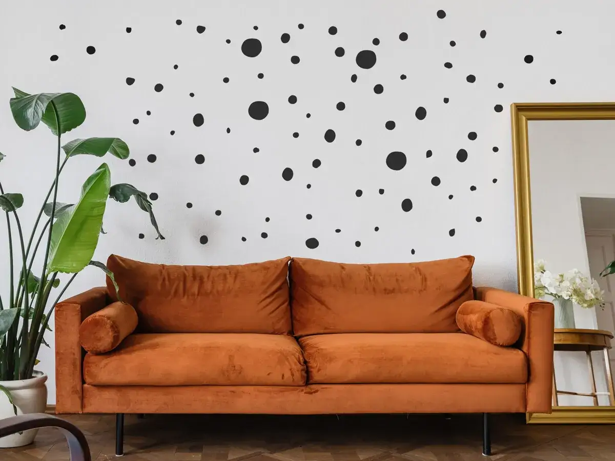 Ein orangefarbenes Samtsofa steht vor einer wei&szlig;en Wand mit vielen schwarzen Polka Dots. Daneben eine gro&szlig;e Pflanze und ein goldener Spiegel.