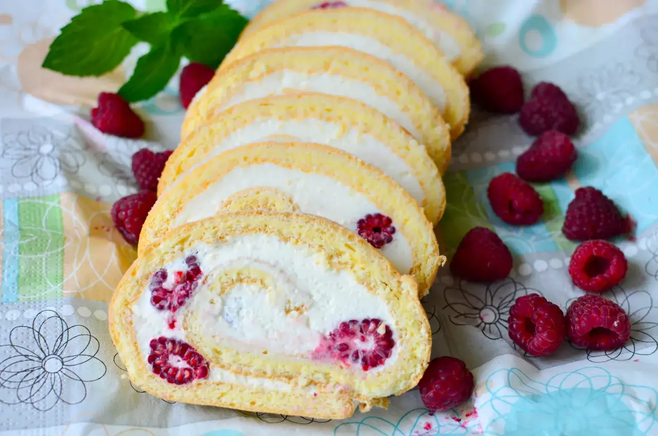 Pyszna rolada biszkoptowa z mascarpone i malinami, pokrojona w plastry, otoczona świeżymi malinami.