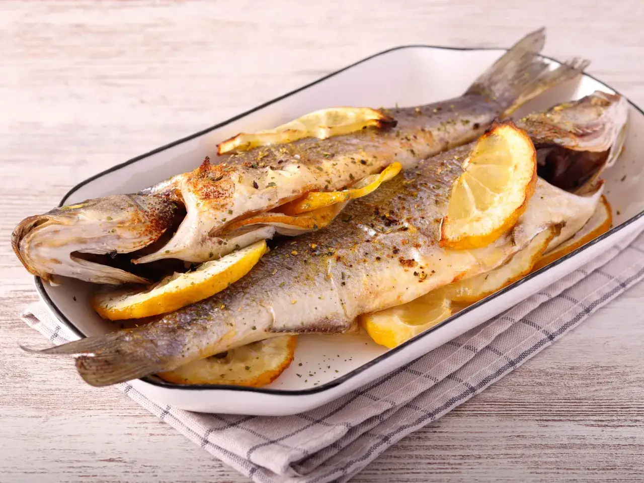 Dos lubinas al horno, doradas y jugosas, servidas con rodajas de limón y hierbas aromáticas en una bandeja blanca.