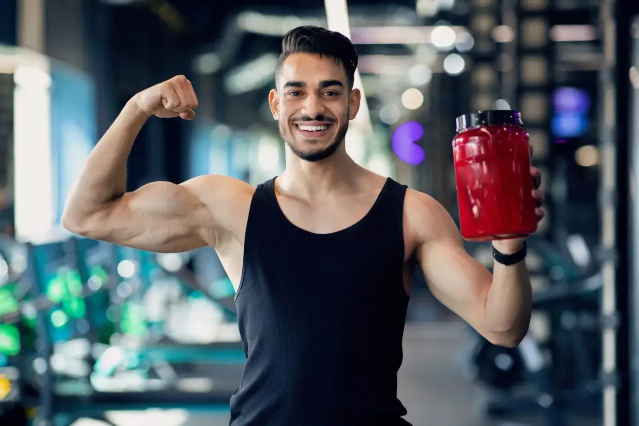Lächelnder Mann im Fitnessstudio zeigt Bizeps und hält eine rote Dose. Er nutzt die BCAA Wirkung für seinen Muskelaufbau.
