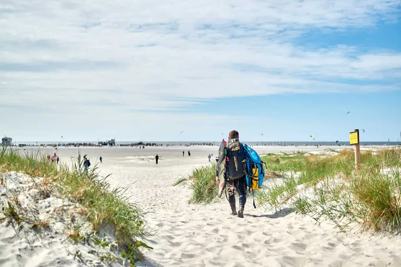 Ein Mann mit Kite-Ausrüstung geht am Strand von St. Peter-Ording Richtung Meer. Im Hintergrund die belebte Innenstadt und viele Kite-Surfer.